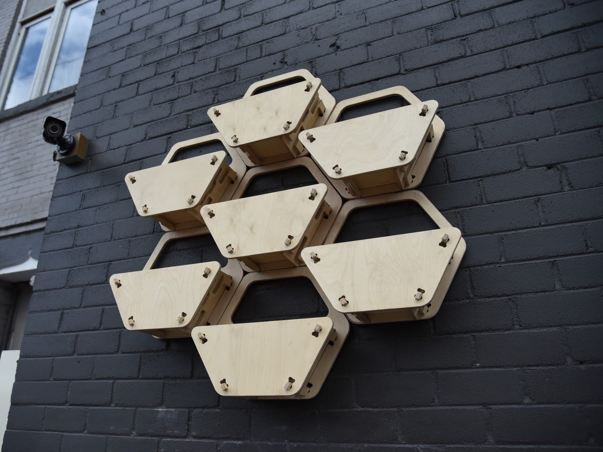 Nine hexagonal plywood planters mounted as a honeycomb array on a dark painted brick wall outside a building