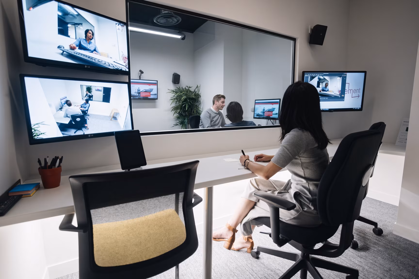 The observation side of a usability lab: a researcher takes notes at a desk while watching a session through one-way glass, with wall monitors carrying the participant's face and screen